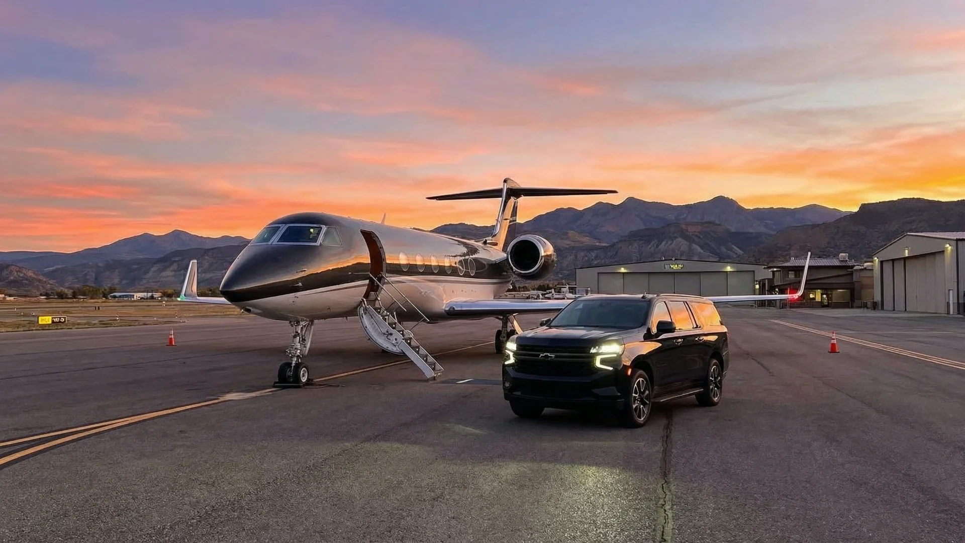 A luxury black Chevrolet Suburban parked on the tarmac next to a private jet at Rifle Garfield County Airport (RIL) during sunset, with the Colorado mountains in the background