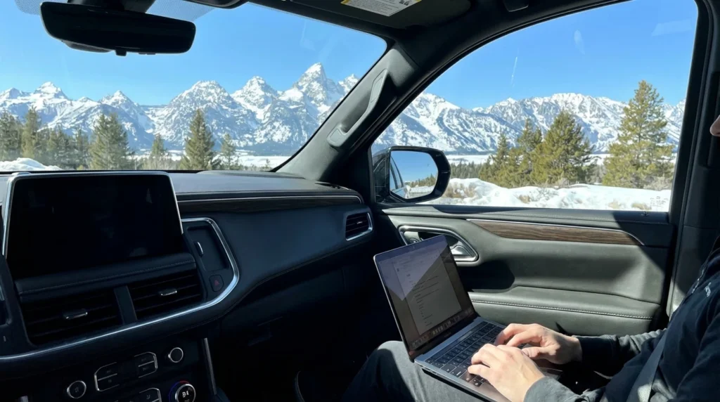 The interior of a luxury SUV showing a passenger working on a laptop with a view of snow-capped mountains through the window.
